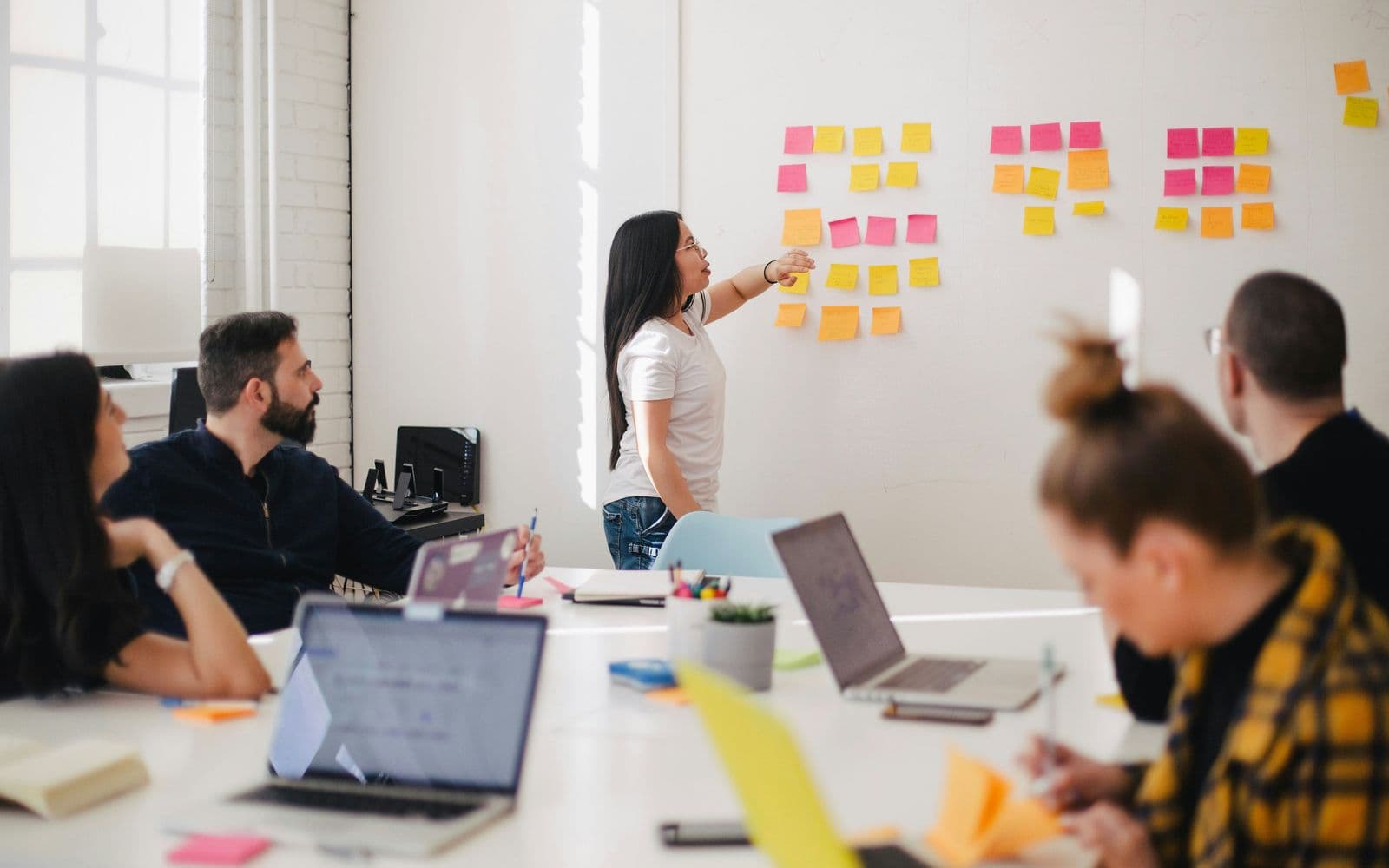 A woman presenting sticky notes on a wall while colleagues sit around a conference table with laptops.